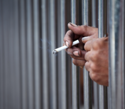 Image of two hands through prison bars, one hand holding a lit cigarette