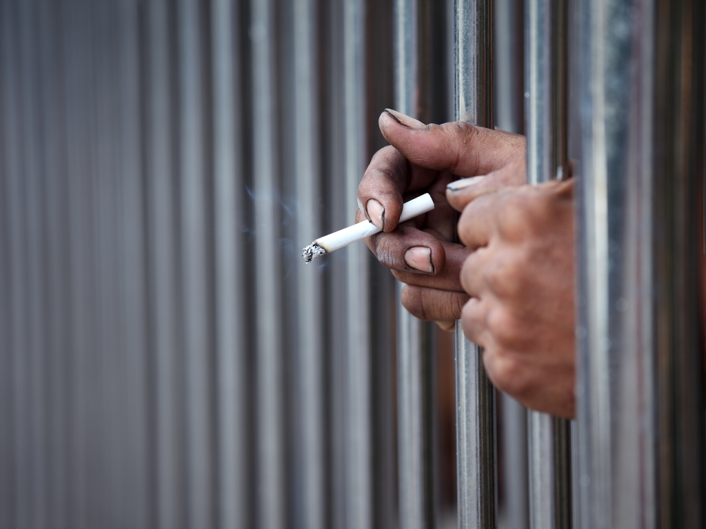 Image of two hands through prison bars, one hand holding a lit cigarette